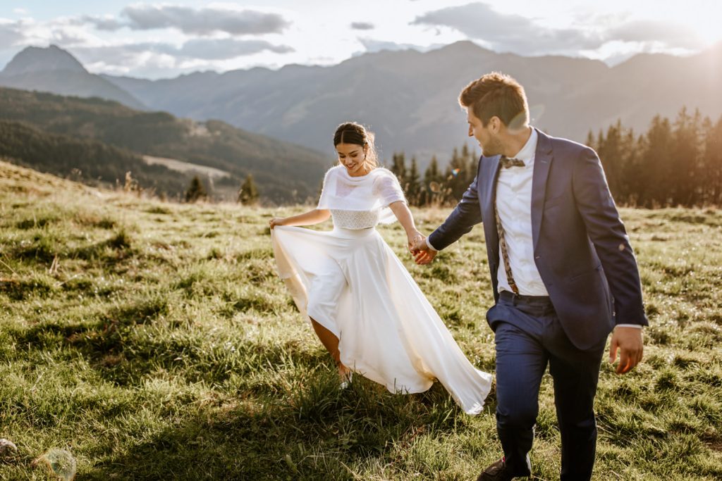 Bräutigam nimmt seine Frau an die Hand und läuft mit ihr auf einer Wiese in Tirol auf den Bergen
