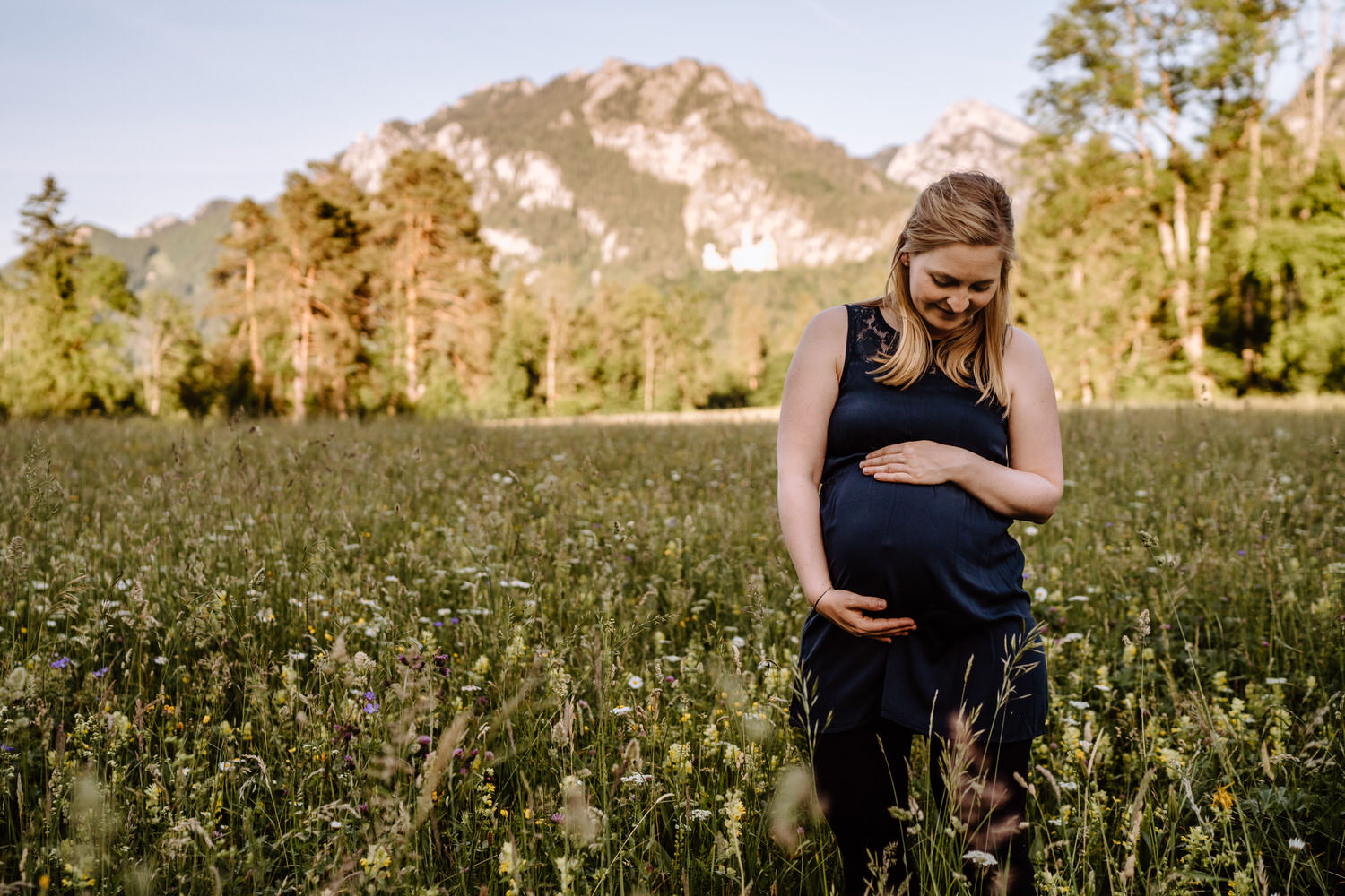 Werdende Mama hält ihren Babybauch auf einer Wiese vor den Bergen am Schwansee